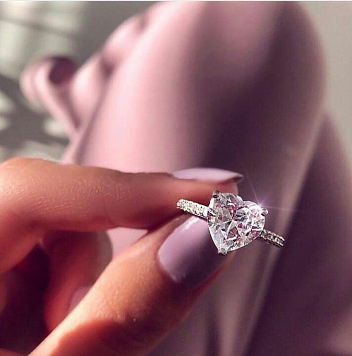 A sparkling heart shaped zircon engagement ring with a silver band adorned with smaller diamonds and white gold plating.