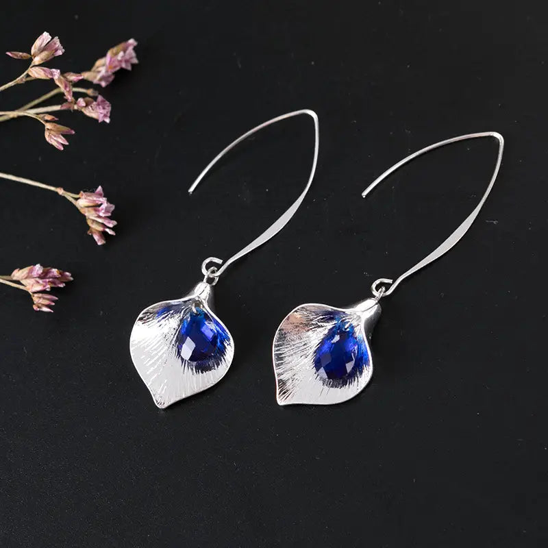 Boucles d’oreilles pendantes en cristal d’orchidée argentée texturée, ornées de formes de feuilles argentées, avec une pierre précieuse facettée bleu profond au centre.