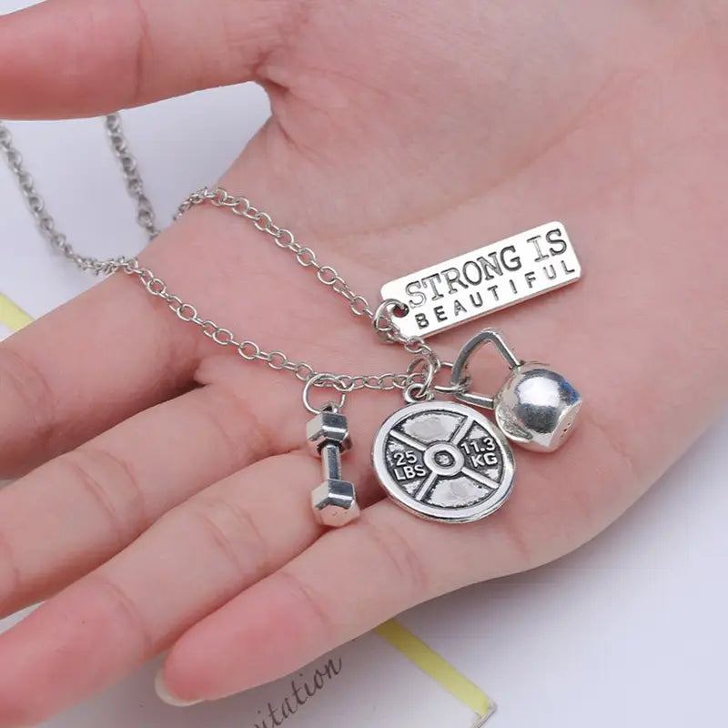 Silver-toned inspirational fitness pendant necklace featuring a rectangular strong is beautiful charm, dumbbell pendant, 25 lbs weight plate, and kettlebell-shaped ball charm.
