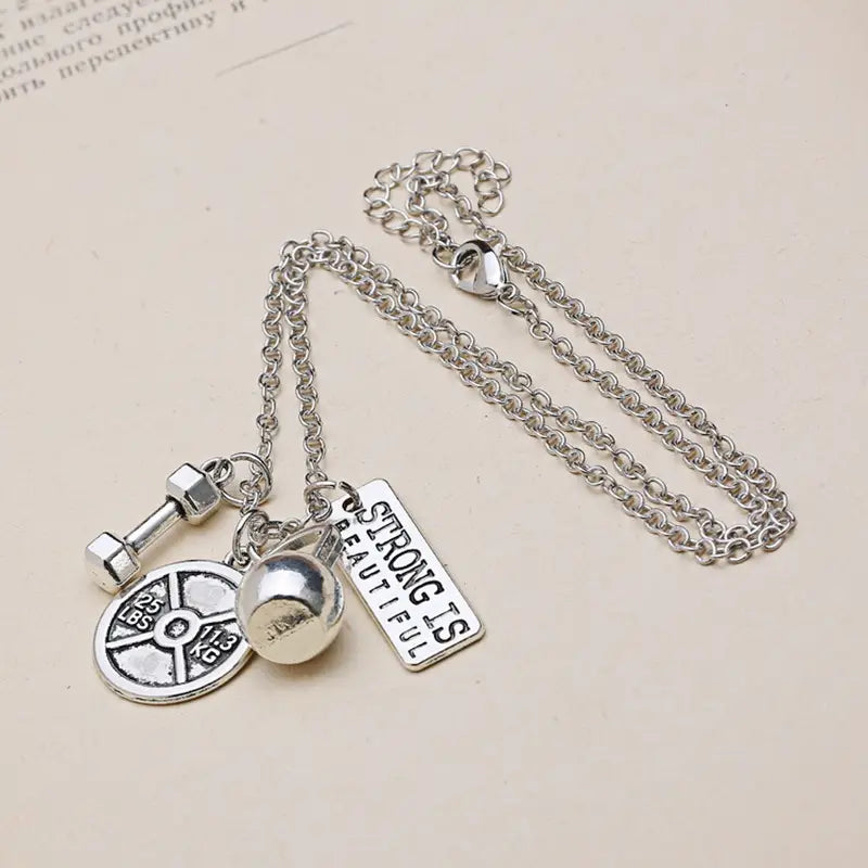 Silver-toned inspirational fitness pendant necklace featuring a chain with multiple gym-themed charms including a dumbbell weight plate and spherical ball.