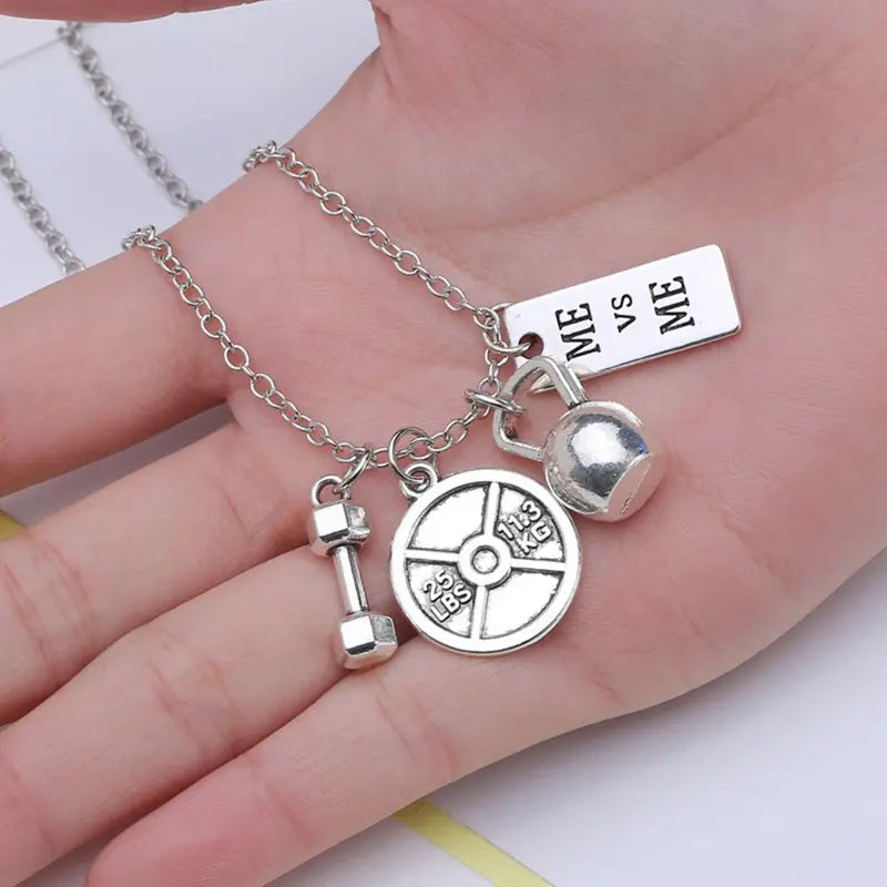 Silver-toned fitness-themed pendant necklace with gym charms including dumbbell weight plate kettlebell and rectangular me vs me tag charm.