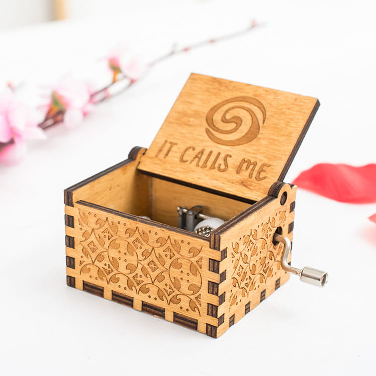 Eine hellbraune hölzerne Musikbox mit Kurbelgriff, mit kunstvoll geschnitzten Motiven, die aus filigranen Blumenschnitzereien bestehen, sowie einem eingravierten Satz, den sie mir über einer Spiralendekoration auf ihrem Deckel zuruft.