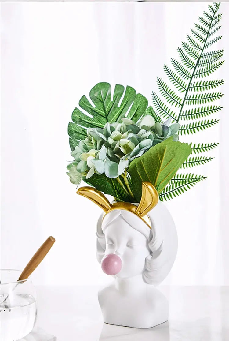 Weiße Keramikvase in Form eines Frauenkopfes mit goldenen hirschgeweihartigen Hörnern und pinkem Kaugummi, der eine Anordnung aus grünen Monstera-Blättern, Farnen und hellblauen Hortensien hält; betitelt „Bubblegum-Girl“ Keramikvase mit goldenen Akzenten.