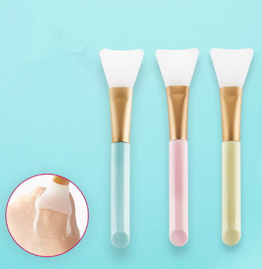 Set of three pastel-colored silicone facial mask brushes with gold-toned iridescent gradient metal handles designed for applying skincare products.