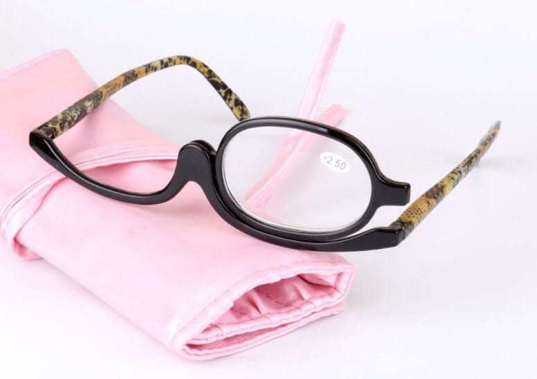 Black-framed makeup reading glasses with camouflage-patterned temples and +2.50 strength lenses resting on a pink fabric case.