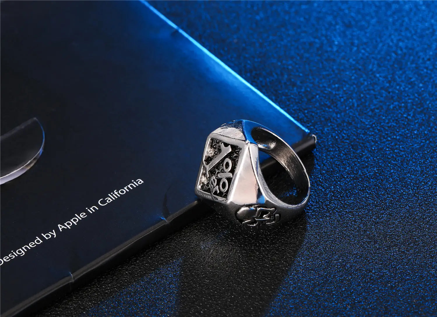 Anillo de sello de biker 1%er de acero inoxidable con acabado plateado, con cara rectangular que presenta letras grabadas y un emblema de calavera en relieve en el lateral, apoyado junto a una superficie azul oscuro
