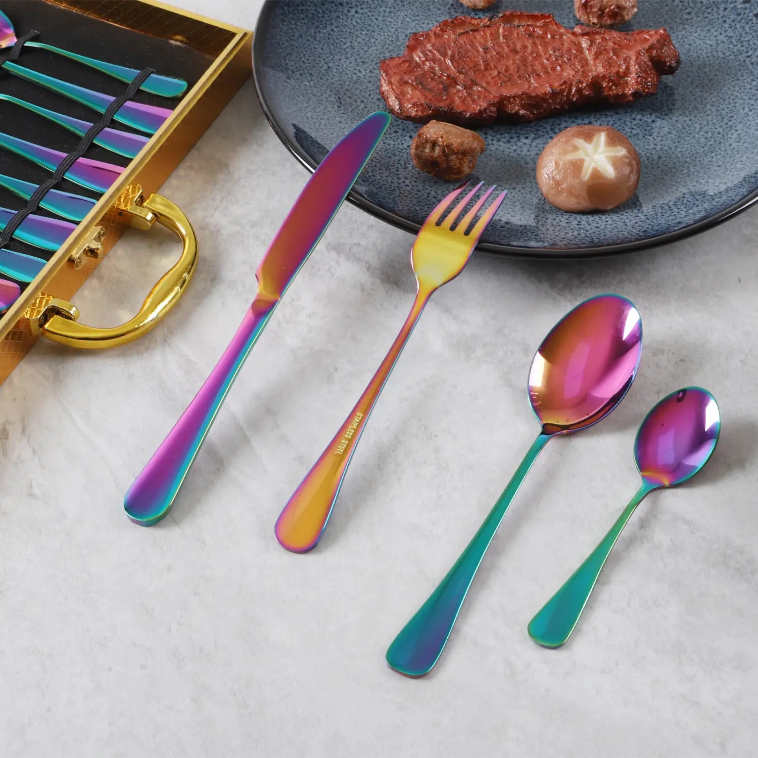 Gold stainless steel cutlery set 24 piece tableware with rainbow-finished iridescent blades and handles displayed alongside a plated steak.