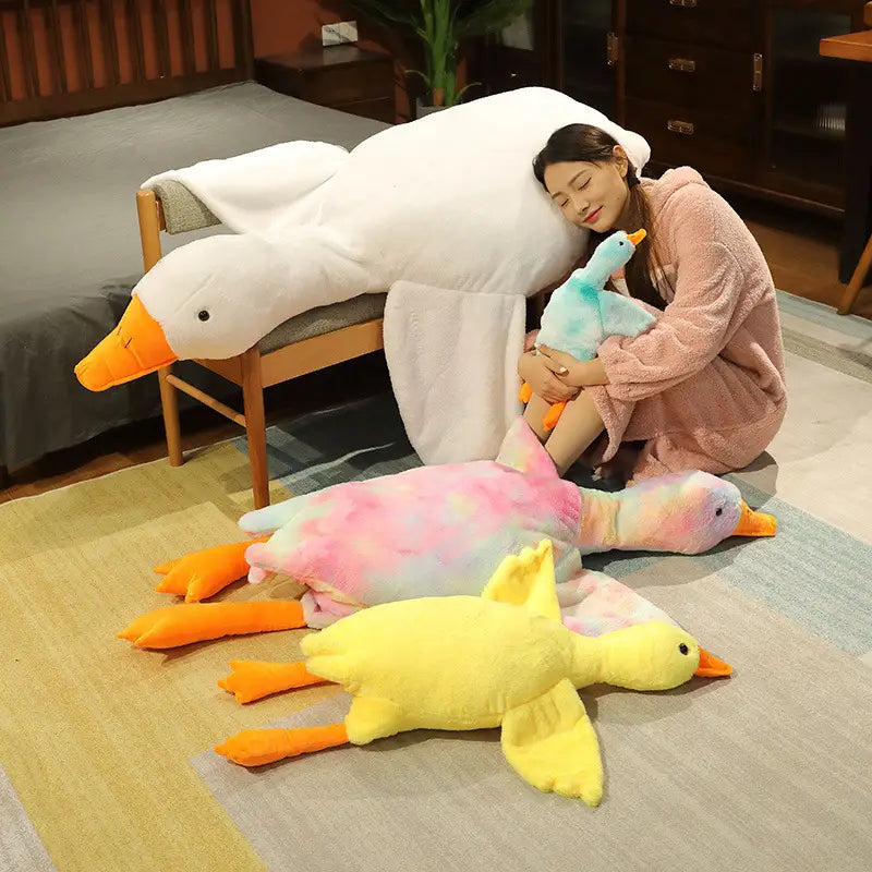 A giant white goose plush sleeping pillow toy with orange beak and feet lying on a bed beside a woman in pink pajamas.