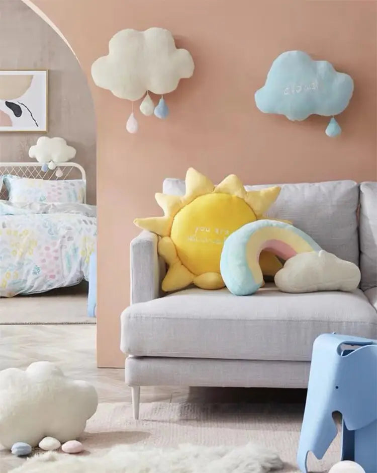 Yellow plush sun pillow with a smiling face and radiating rays resting on a gray sofa for cloud sun rainbow plush wall hanging decor.