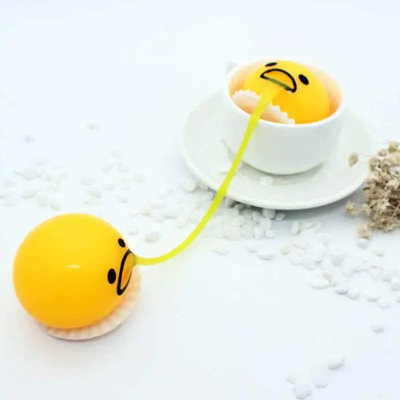 Yellow Gudetama egg-shaped stress ball with a smiling face connected by a yellow elastic band to an identical figure sitting in a white teacup used as a vomiting egg yolk stress relief toy.