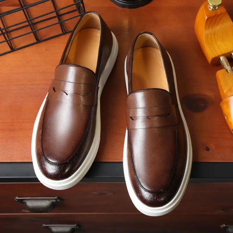 Mocasines de cuero marrón estilo británico para hombres con suelas de goma blancas y detalles de costura.