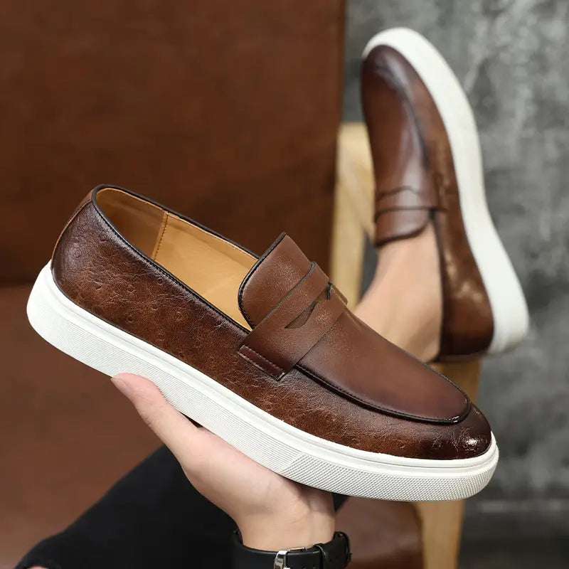 Mocasines tipo slip-on para hombres estilo británico de cuero marrón con suela de goma blanca y textura sutil.