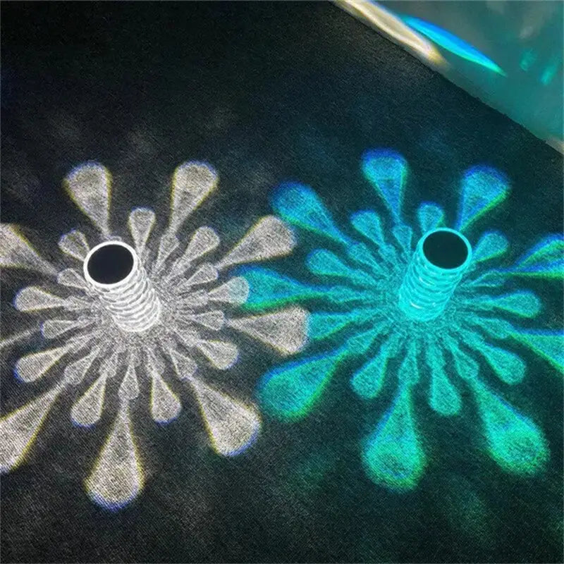 Two glowing flower-like patterns in silver and teal with radiating petals projected onto dark fabric by a crystal table lamp with touch control diamond LED night light.
