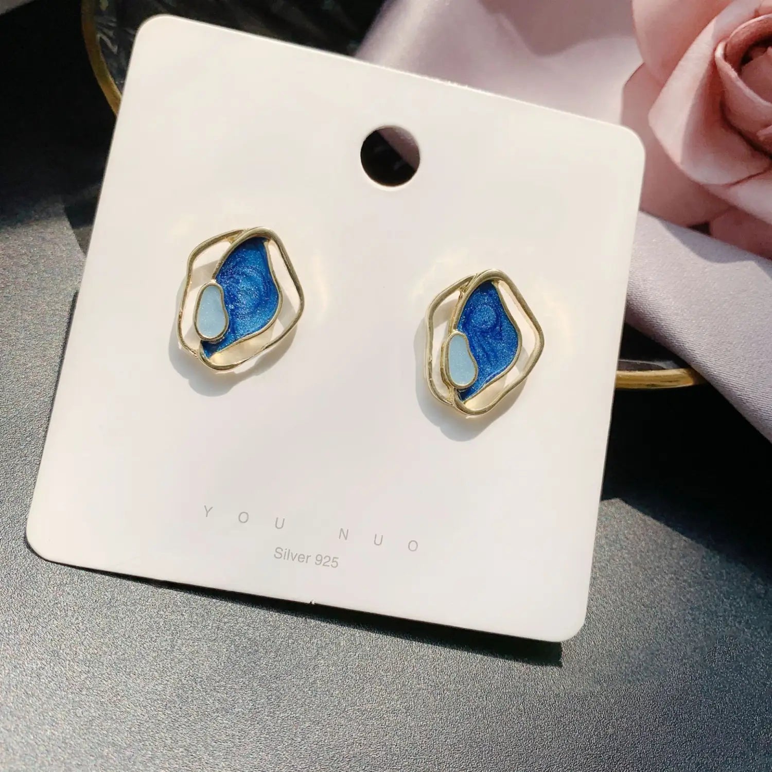 Une paire de boucles d’oreilles clous géométriques élégantes en émail bleu et blanc, en argent géométrique, montées dans un cadre métallique de ton doré et présentées sur une carte de marque.