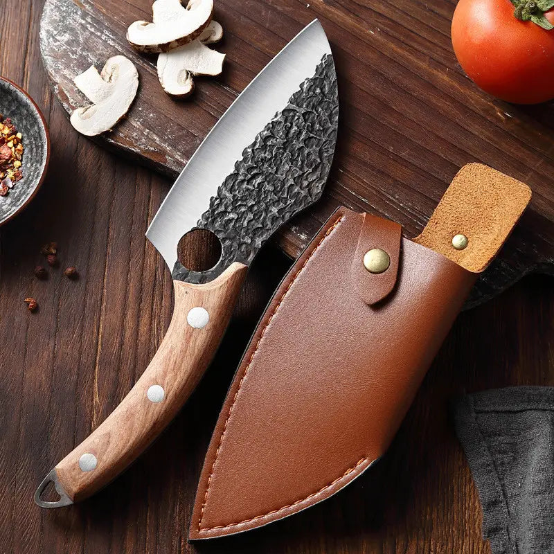 Un couteau de cuisinier d’extérieur forgé en acier à haute teneur en carbone, au style rustique, avec une lame en acier noirci martelée, un manche en bois clair fixé par des rivets argentés et un étui en cuir brun.