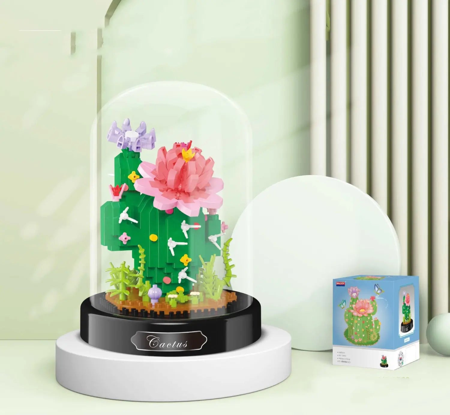 Un ramillete de piezas de construcción florales en cúpula de vidrio que presenta una escultura de cactus verde hecha con ladrillos de plástico entrelazados, con una flor rosa grande y pequeños pájaros blancos.