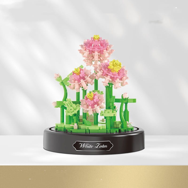 un ramillete de piezas de construcción florales en cúpula de vidrio que presenta una delicada disposición de flor de loto rosa y amarilla elaborada con ladrillos de plástico verdes entrelazados, mostrada sobre una base circular negra con un loto blanco grabado en escritura plateada