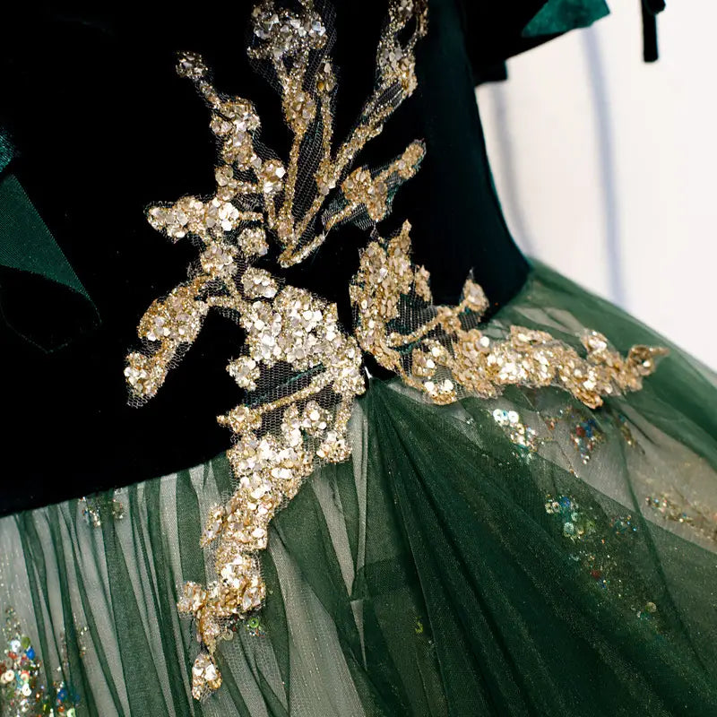 Robe émeraude en velours à épaules dénudées, ornée d’une applique dorée irisée en forme de feuille, rehaussée de cristaux scintillants et de sequins sur un corsage riche en velours vert émeraude.