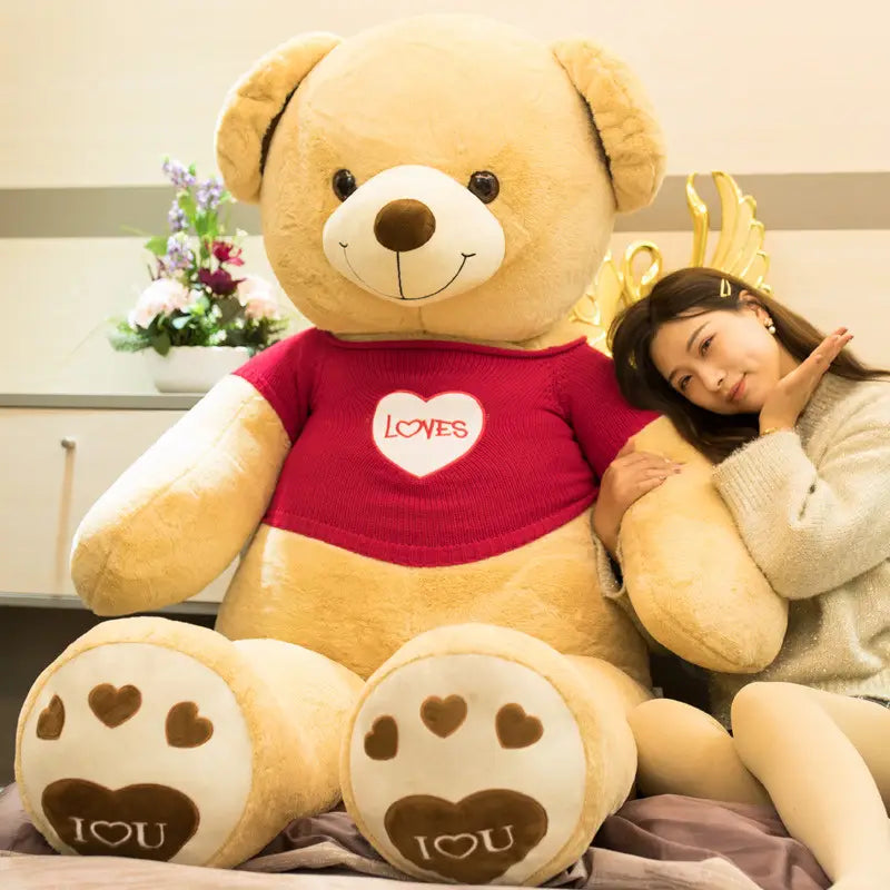 Ein riesiger beigefarbener Plüsch-Teddybär, der einen roten gestrickten Pullover mit Herzmotiven trägt, mit dem Text „loves“ sowie braunen Herz-Pfotenpolstern mit einer „I heart u“-Nachricht.