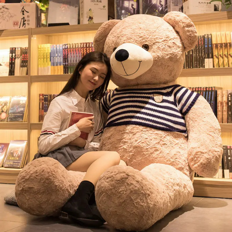 Ein Plüsch-Groß-Teddybär-Spielzeug in hellbrauner Farbe, der einen dunkelblau-weiß gestreiften Strickherzpullover trägt.