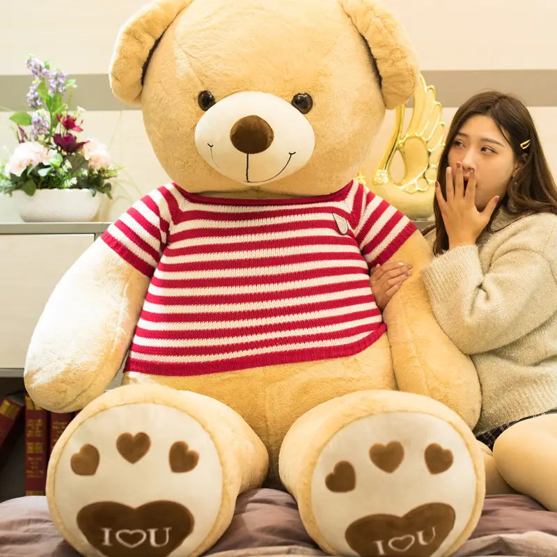 Ein riesiger beigefarbener Plüsch-Teddybär, der ein rot-weiß gestreiftes Strickhemd mit herzgemusterten Pfotenpolstern trägt, auf denen „i heart u“ steht.