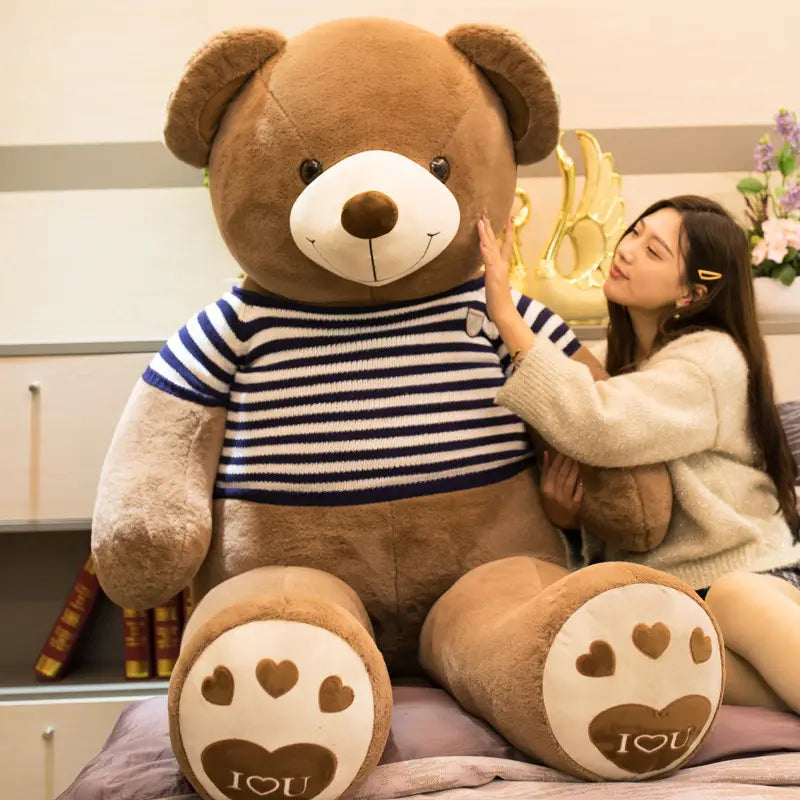 Ein riesiger brauner Plüsch-Teddybär, Plüschspielzeug, der einen dunkelblau-weiß gestreiften Pullover trägt, mit Herzdesigns auf seinen Pfoten.