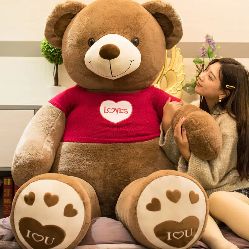 Ein riesiger brauner Plüsch-Teddybär-Plüsch, der einen roten Pullover trägt, mit einem weißen Herz, auf dem „loves“ steht, sowie Pfotenabdrücken an seinen Füßen.