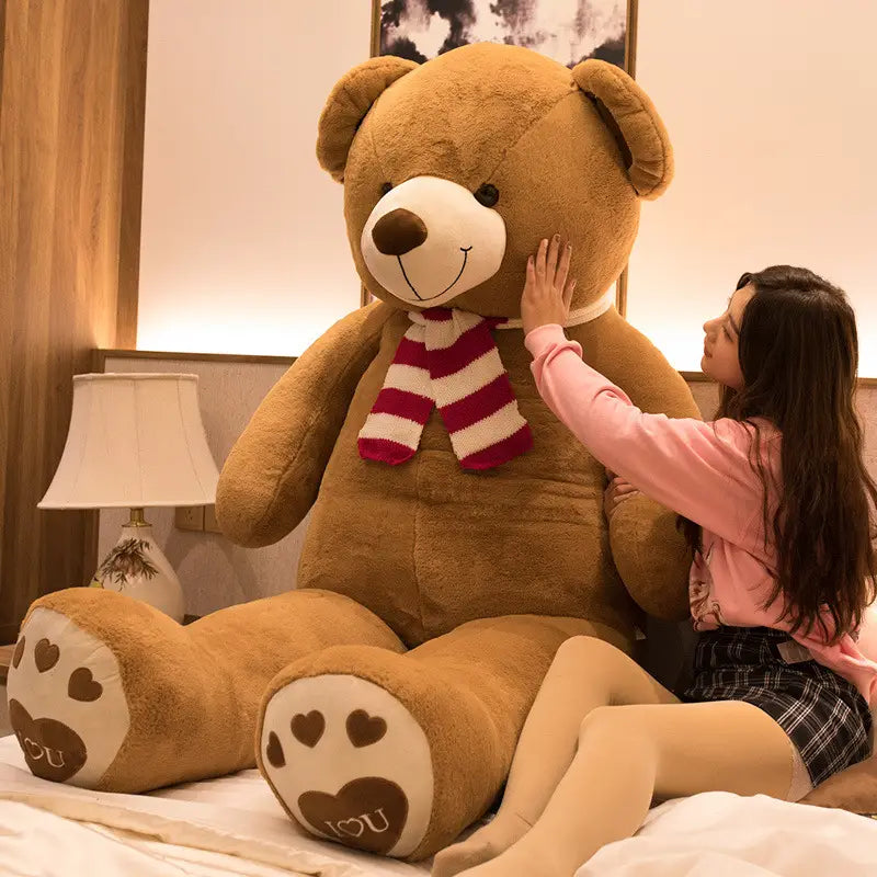 Ein riesiger brauner Plüsch-Teddybär mit weißem Schnäuzchen und brauner Nase trägt einen rot-weiß gestreiften Schal und herzgemusterte Pfotenpolster, die das Wort „you“ tragen.