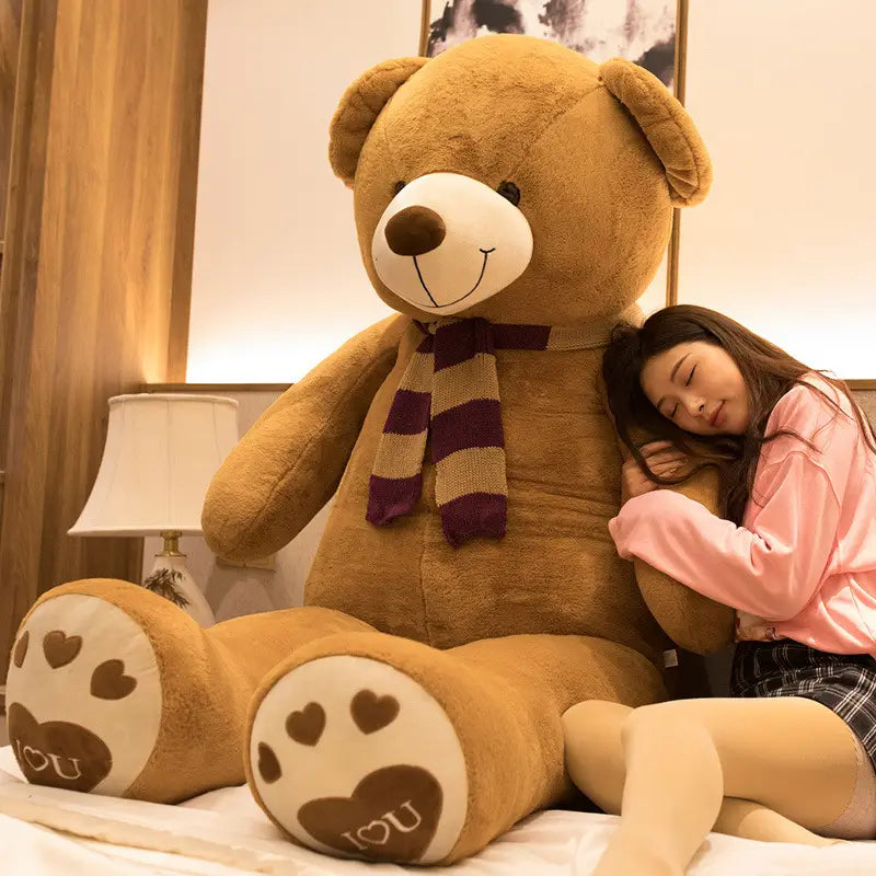 Ein riesiger Teddybär-Plüsch in beiger Farbe mit weißem Schnäuzchen und brauner Nase, der einen gestreiften Schal trägt und Pfotenabdrücke zeigt, auf denen „i love you“ steht.