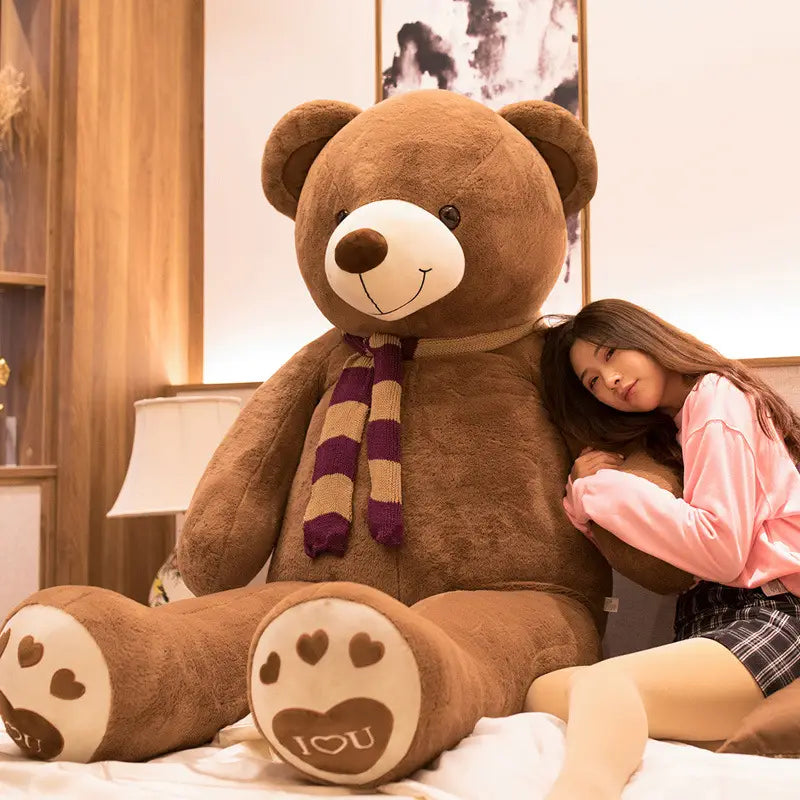 Ein riesiger brauner Plüsch-Teddybär mit weißem Schnäuzchen, schwarzen Knopfaugen und Pfotenpolstern mit Herzdesigns sowie dem Spruch „i love you“, der einen lila- und beigen gestreiften Schal trägt.