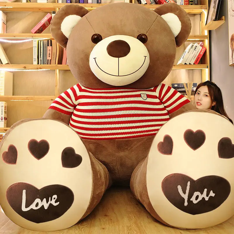 Ein großer brauner Plüsch-Teddybär, der einen roten und weißen gestreiften Herzpullover trägt, mit „love“ und „you“, die auf seinen Pfoten geschrieben stehen.