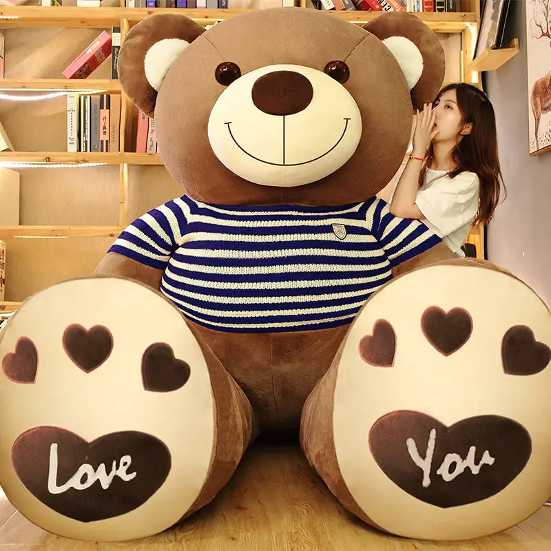 Ein riesiger brauner Plüsch-Teddybär, der ein blau-weiß gestreiftes Strickhemd trägt, mit „love“ und „you“, die auf seinen Pfoten bestickt sind.