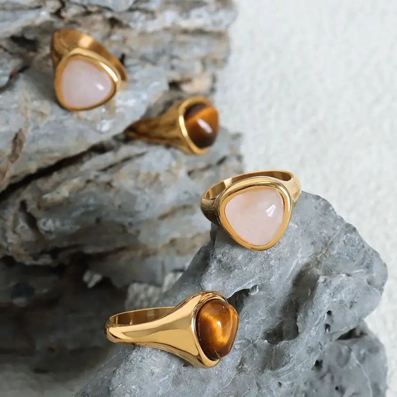 Gold-toned heart shaped natural stone titanium steel signet rings featuring smooth triangular-cut rose quartz and tiger eye stones set in sleek polished metal bands.