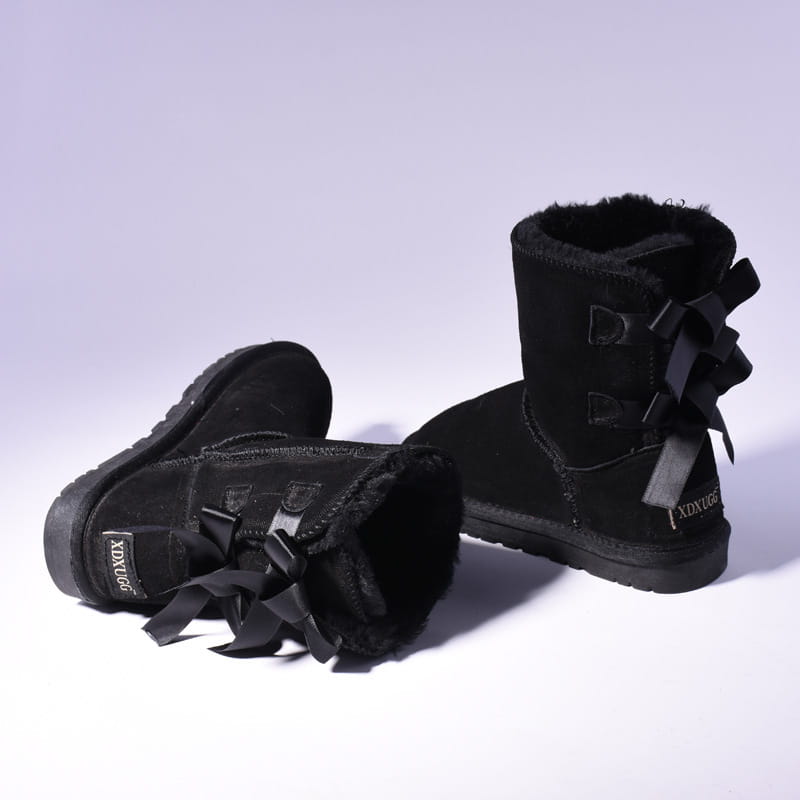 Black ugg-style suede ankle boots with plush shearling lining and decorative bows on the outer side designed for winter wear.