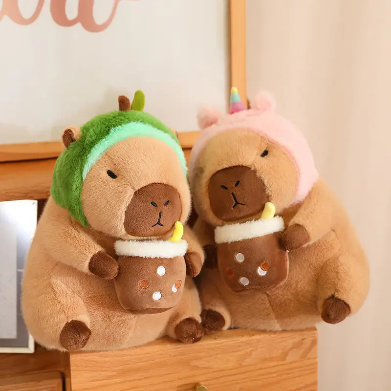 Two plush capybara toys featuring a green headband and a rainbow horn with pink ears, each holding a brown cup accessory, dressed in bread and flower outfits.