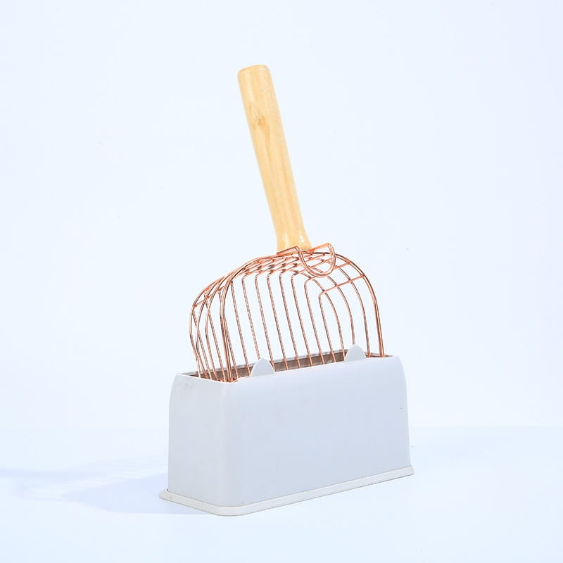 Pala para arena sanitaria de gato de metal color rosa dorado de diseño moderno y elegante, con malla de alambre y mango de madera, colocada en su base de plástico blanco a juego.