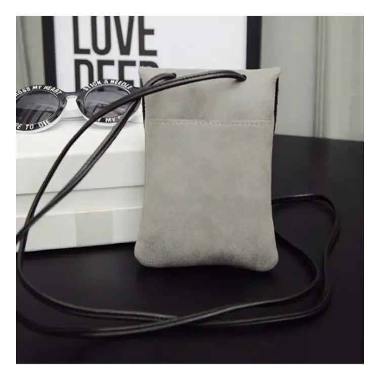 A sleek light gray matte suede vegan leather crossbody pouch with a black adjustable strap and subtle stitching details from the old worldly charm collection.