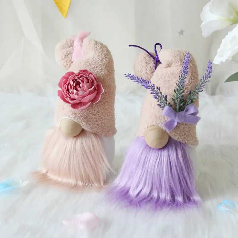 Two plush beige-furred gnome dolls with fluffy pink and purple beards adorned with decorative flowers and lavender sprigs displayed as desktop ornaments.