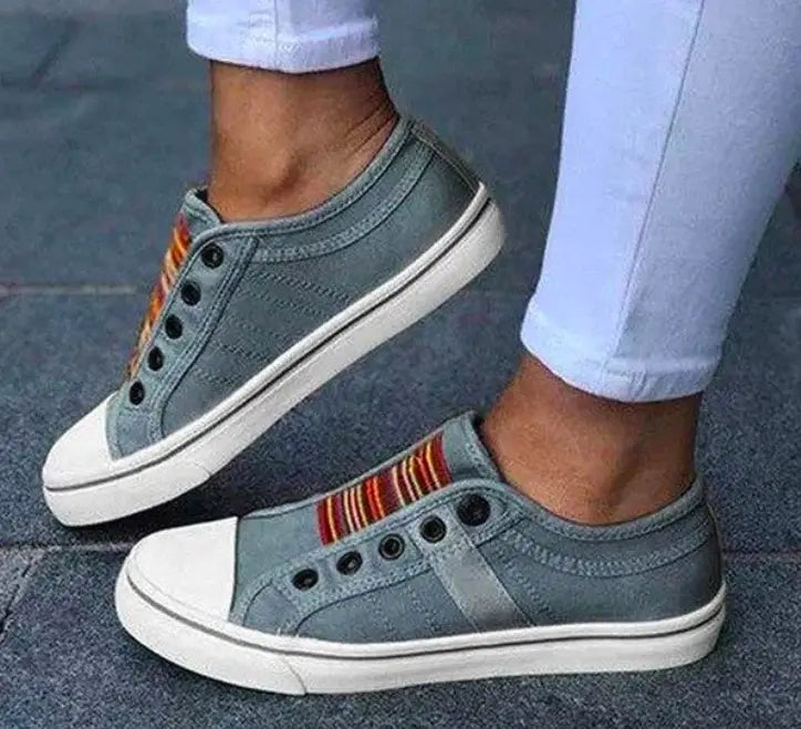 Graue schnürlose Canvas-Sneaker mit weißen Gummisohlen und einem farbigen gestreiften Akzent entlang der Seite, gestaltet als Loafer zum Hineinschlüpfen.