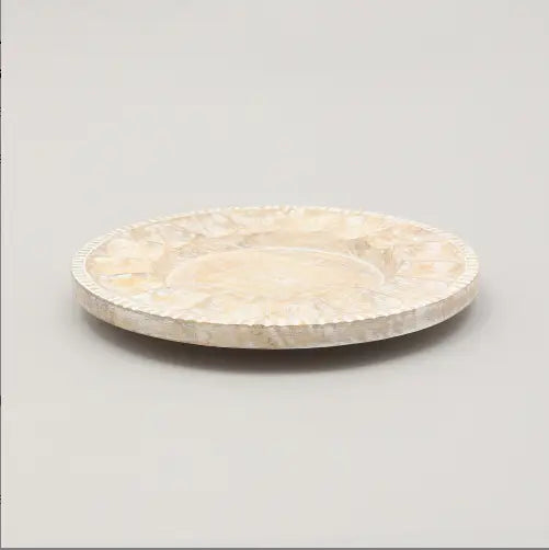 Une assiette plate délicate en bois nacré blanc cassé, avec un bord festonné et un motif d’incrustation florale subtile, provenant d’un ensemble de table rustique fait à la main.