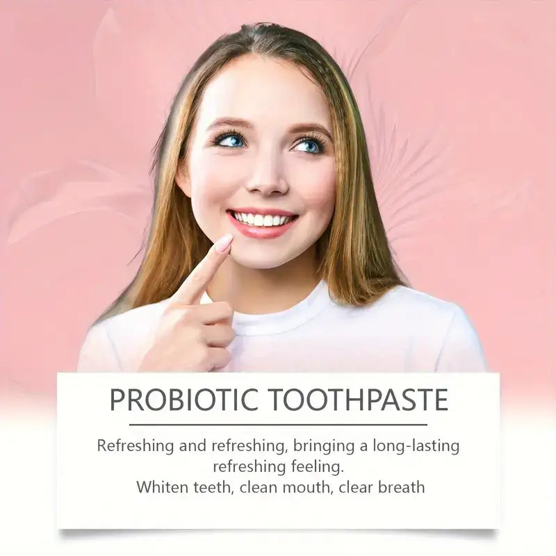 A woman with light brown hair and blue eyes wearing a white top smiles while pointing to her teeth against a soft pink background promoting probiotic toothpaste for deep cleaning and fresh breath.