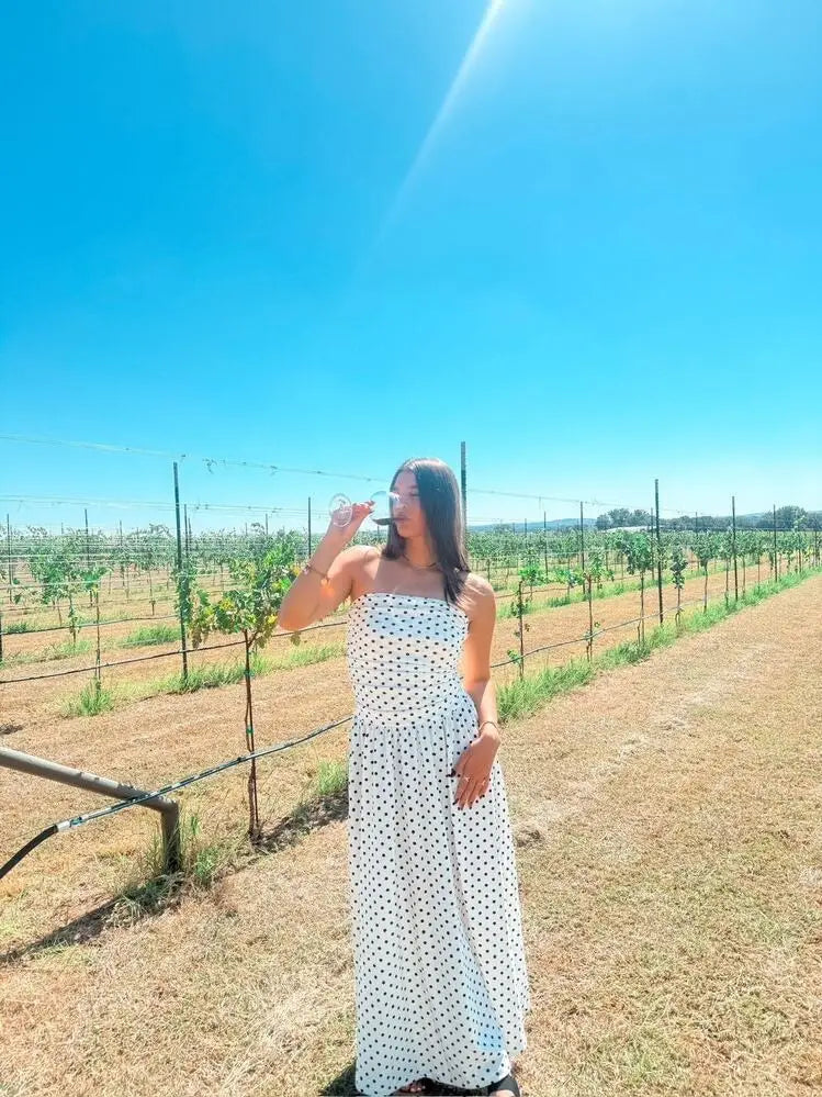 A woman wearing a white polka dot off shoulder strapless maxi dress stands outdoors sipping from a clear wine glass.