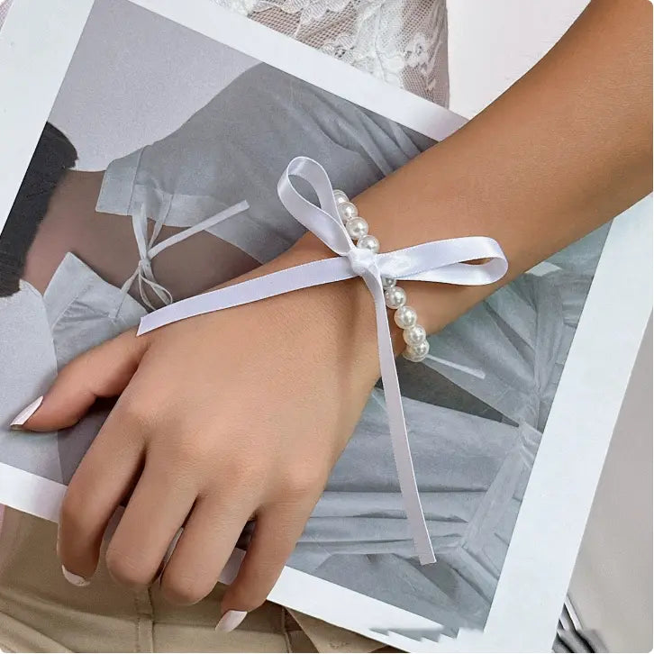 A delicate white satin ribbon bracelet adorned with a row of small lustrous pearls and tied into an elegant bow, part of a black satin and pearl ribbon choker bow jewelry set.