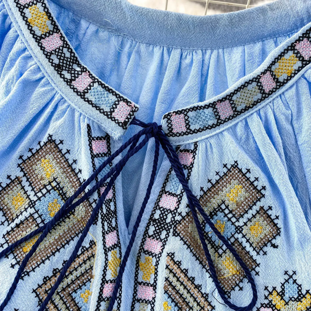 Light blue embroidered lantern sleeve tie neck blouse with intricate pink yellow and brown cross-stitch patterns on soft cotton fabric.