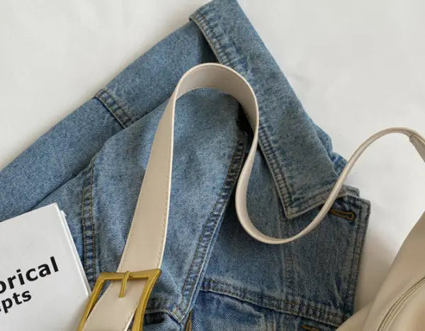 Cream-colored leather shoulder bag with gold buckle and strap displayed with a matching leather belt with gold-toned rectangular buckle on a folded denim jacket.