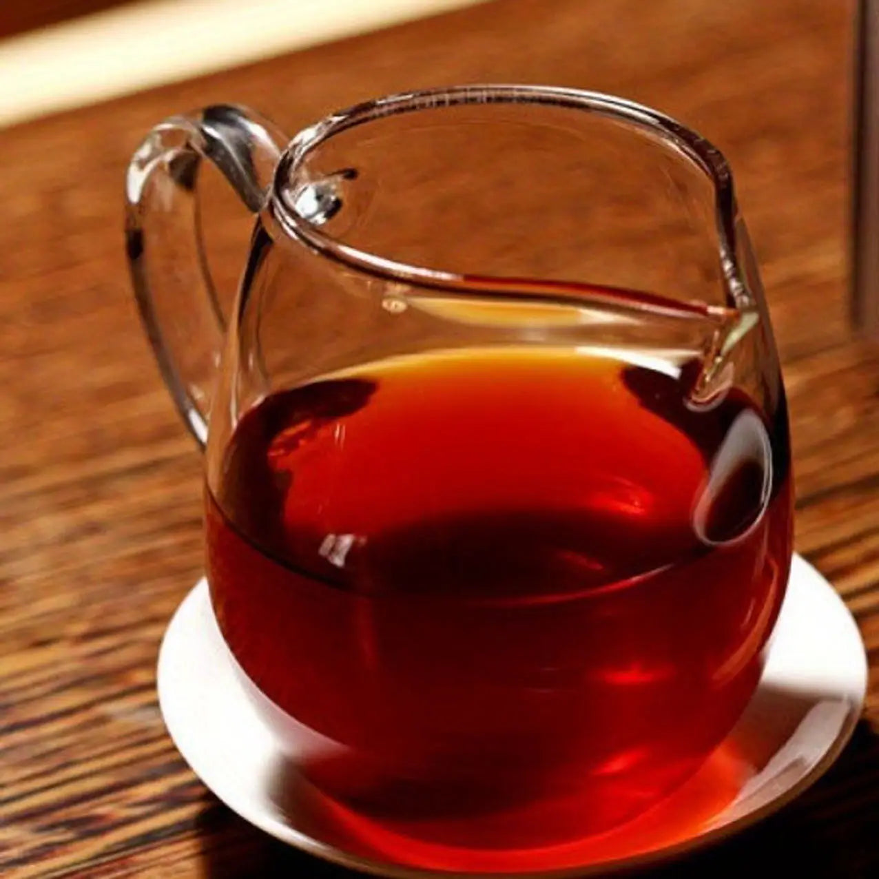 Eine klare Glas-Karaffe, gefüllt mit reichhaltigem bernsteinfarbenem Premium-Yunnan-Reif-Puer-Tee, der auf einer weißen Untertasse ruht.