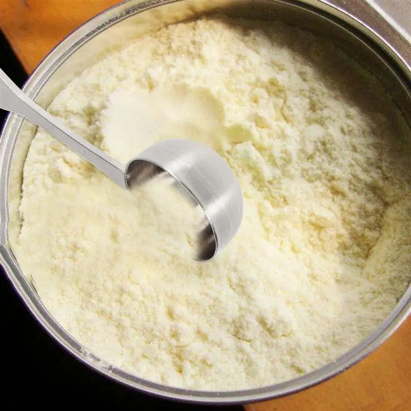 A stainless steel 304 measuring spoon with a polished finish resting in a metal container filled with creamy white powdered milk.