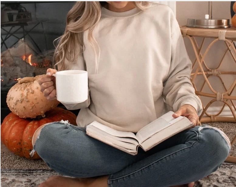 Taza de cerámica blanca sostenida en la mano junto con una sudadera tipo crewneck de algodón oversize en color beige.
