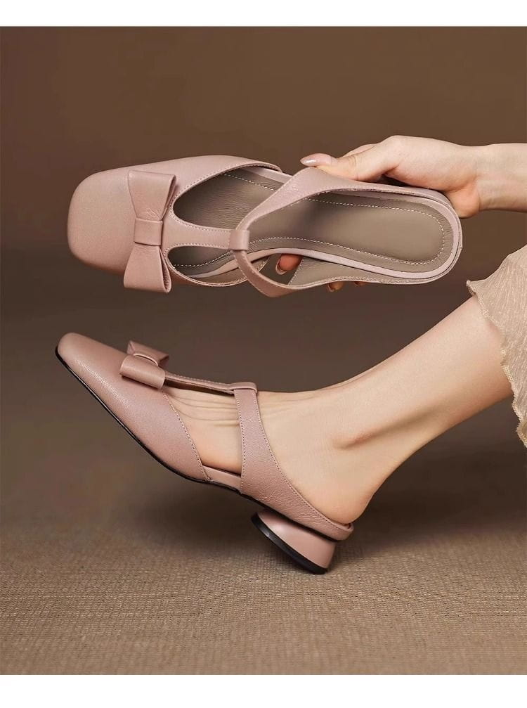 A pair of blush pink leather mule flats for women featuring a closed toe, t-strap design, and a small bow on the front.