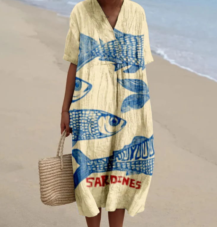 A flowing pale yellow maxi dress with a v-neck and short sleeves features bold blue fish patterns and red lettering spelling sardines on the front.